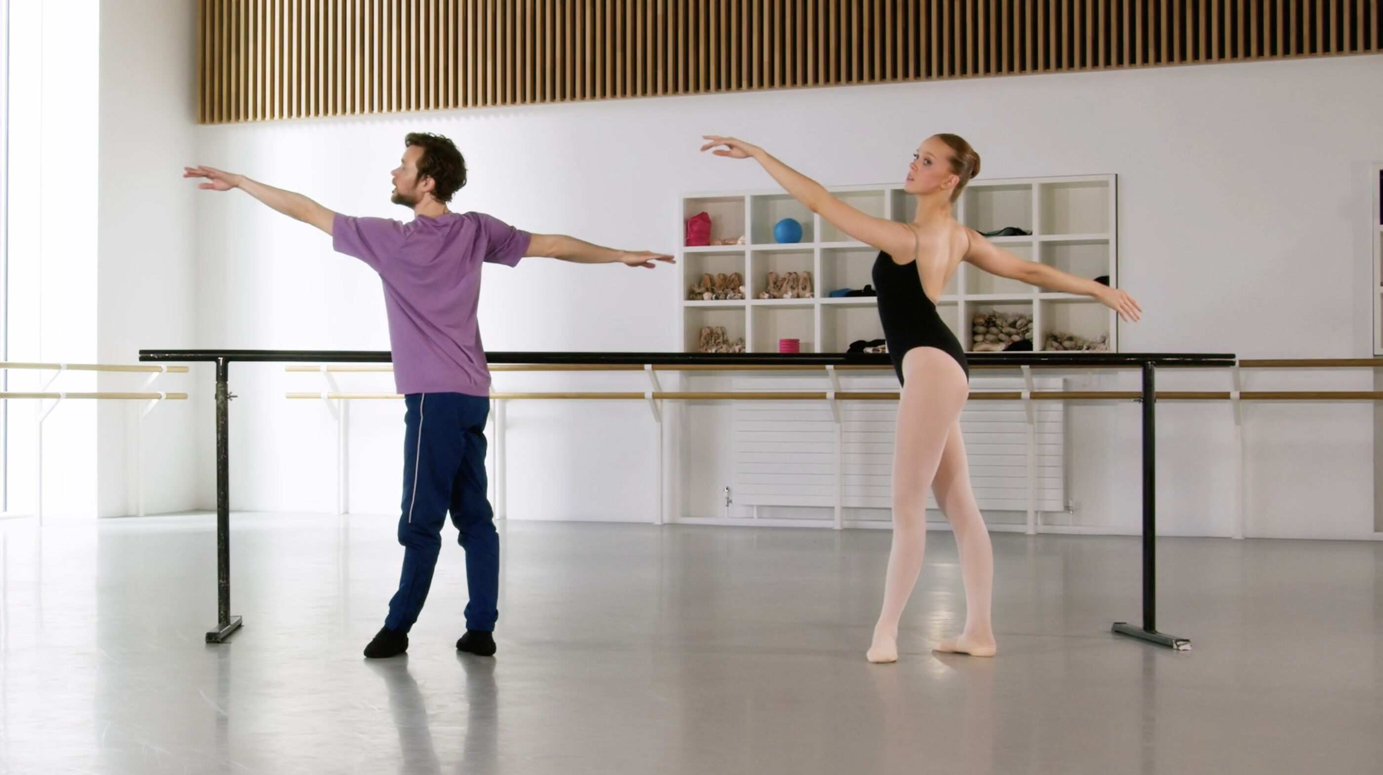 A ballet tutor and a demonstrator in an arabesque position at the barre