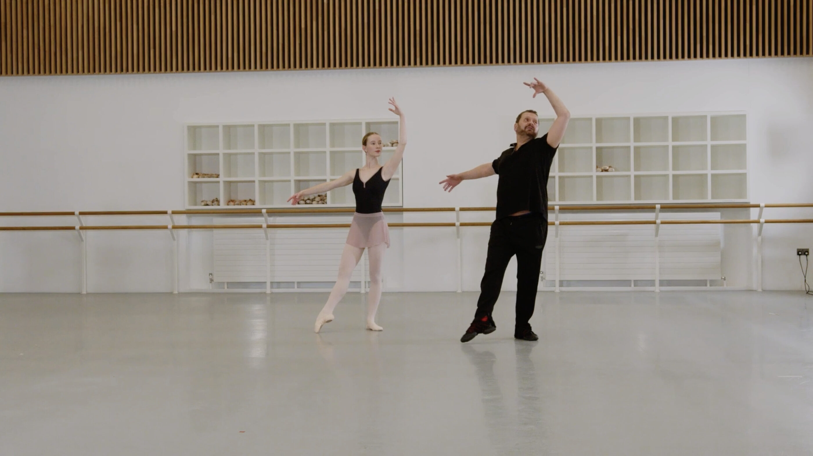 A ballet teacher and a demonstrator are doing a tendu en croisé in a dance studio.