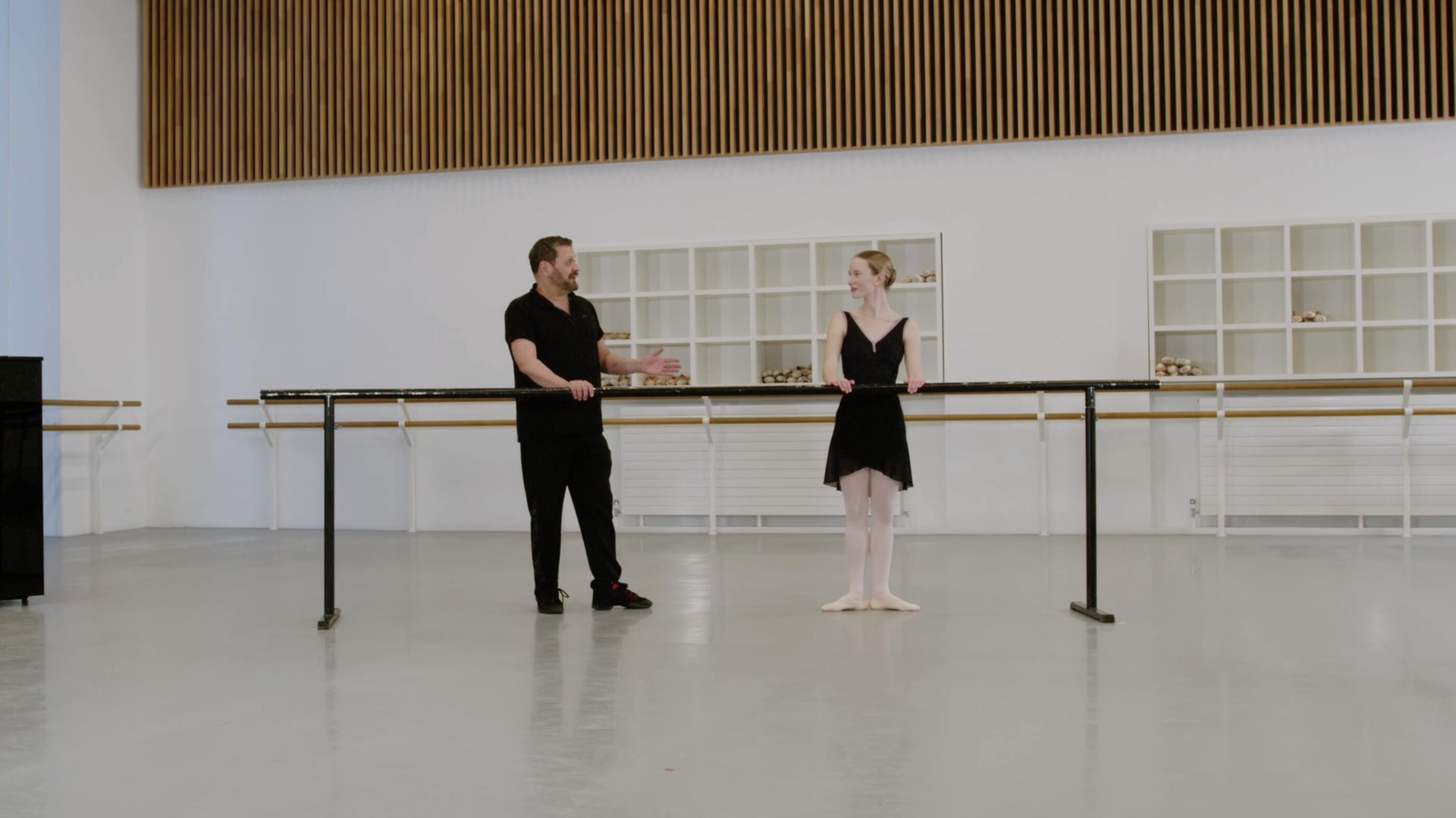 A ballet teacher and a demonstrator are standing at the barre in a dance studio. The teacher gestures towards the demonstrator who's standing in first position.