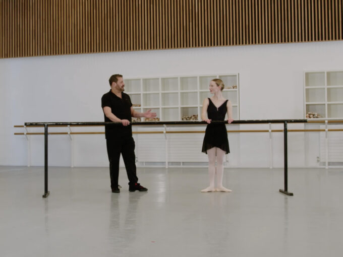 A ballet teacher and a demonstrator are standing at the barre in a dance studio. The teacher gestures towards the demonstrator who's standing in first position.