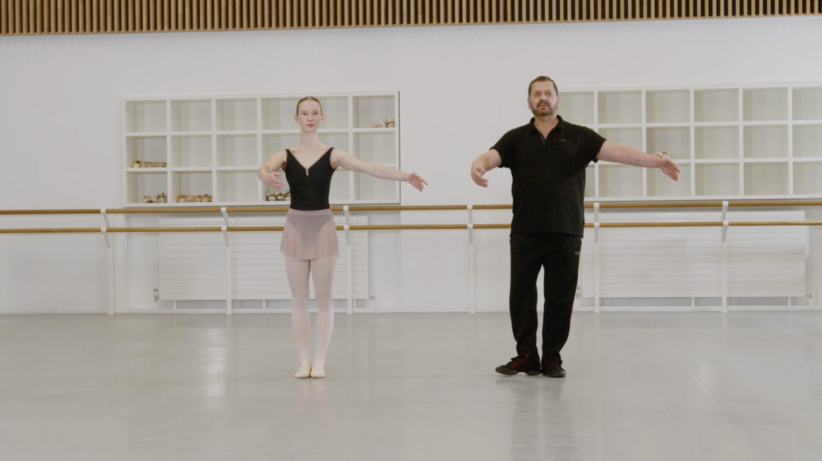 A ballet teacher and a demonstrator are showing the arms preparation for a turn. The teacher is wearing a black t-shirt and trousers. The demonstrator is wearing a black leotard and a pink skirt.