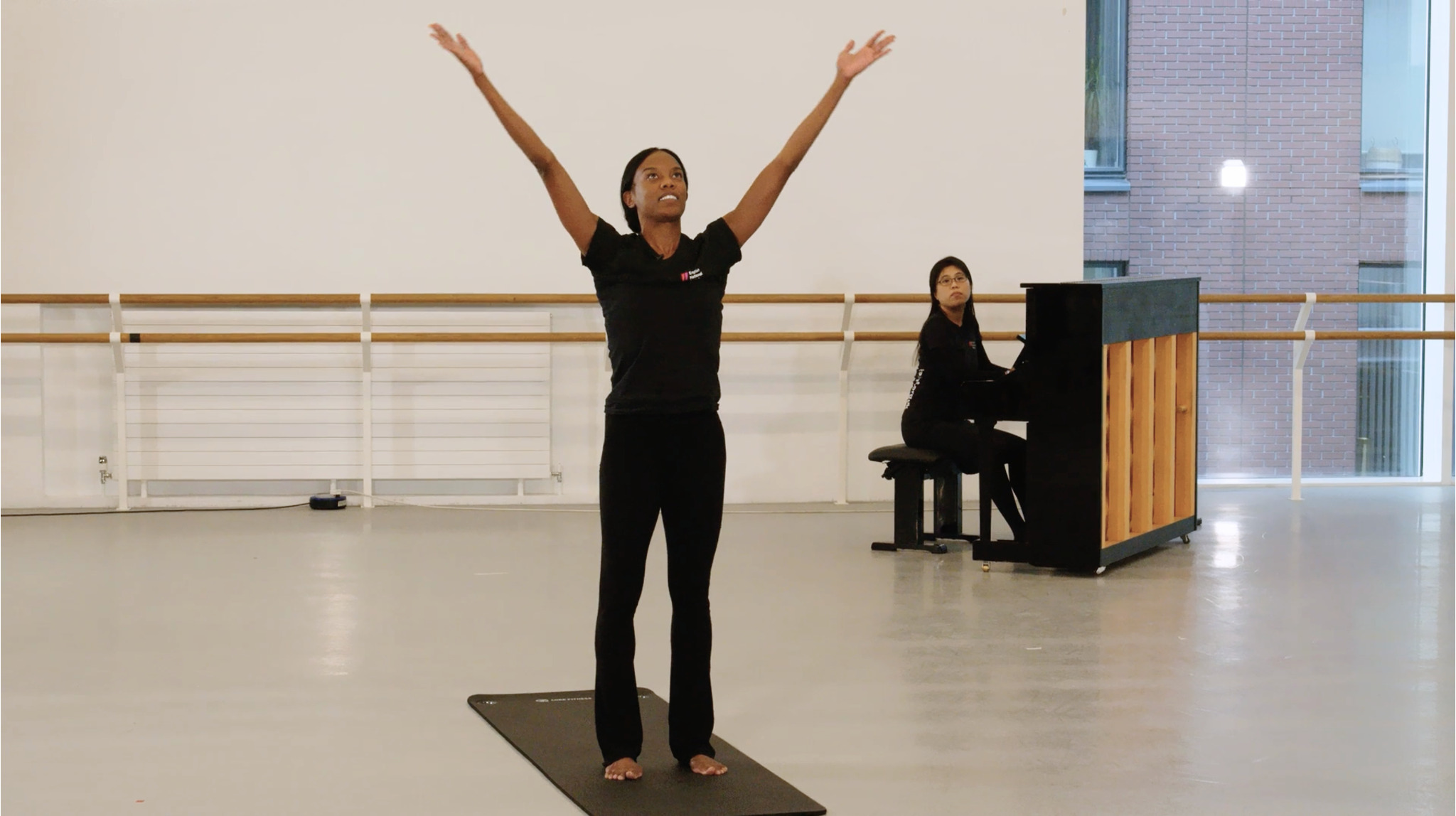 A woman in black attire stands on a yoga mat, her arms lifted towards the ceiling