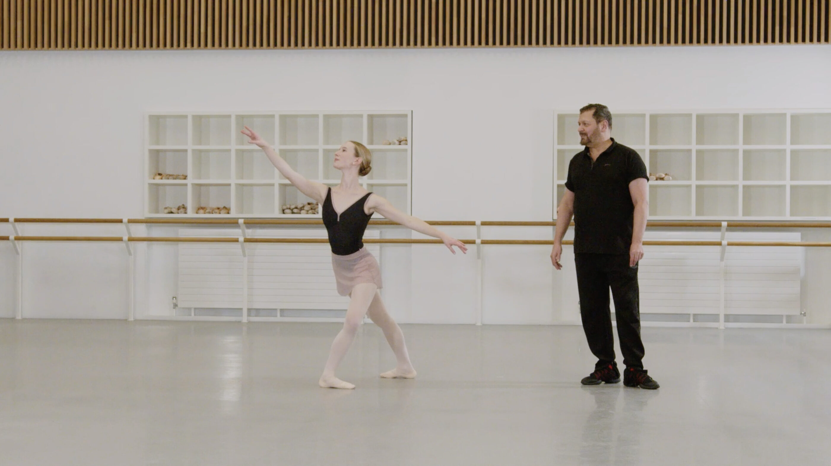A ballet teacher and a demonstrator are in a studio. The demonstrator has just finished a pirouette. The demonstrator is wearing a black leotard and pink skirt. The teacher is wearing black clothing.