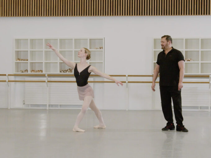 A ballet teacher and a demonstrator are in a studio. The demonstrator has just finished a pirouette. The demonstrator is wearing a black leotard and pink skirt. The teacher is wearing black clothing.