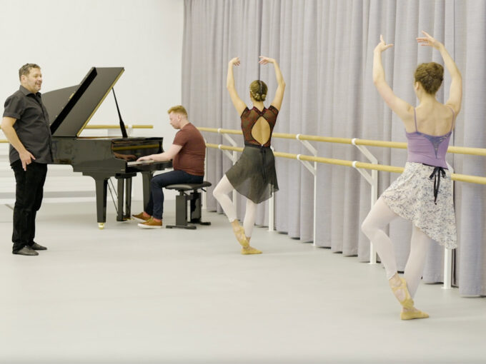 Two dance demonstrator are at the barre, in cou de pie position. A dance instructor is on the side of the screen, while a pianist is in the background.