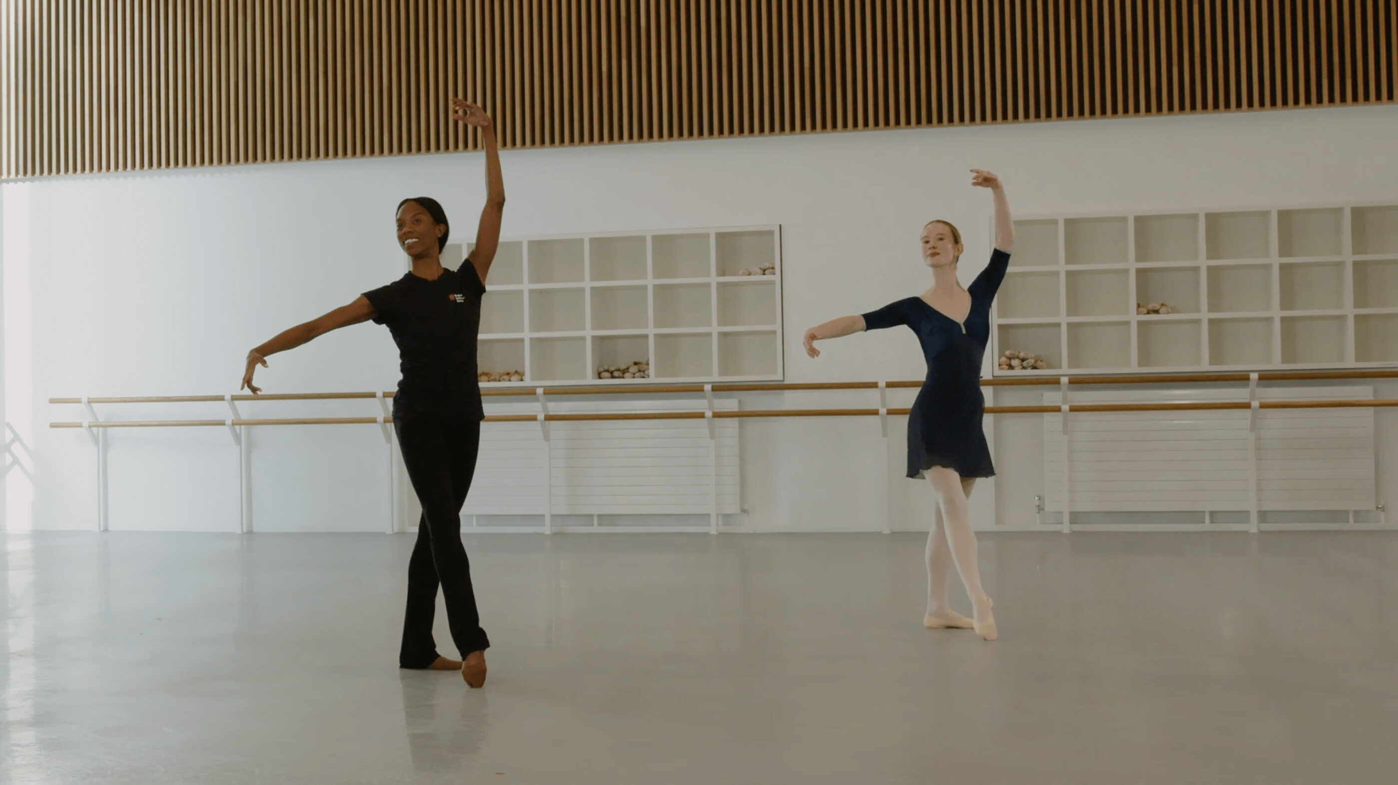 A ballet teacher and a ballet demonstrator are doing a battement tendu at the centre in a studio. The teacher is wearing a black outfit, the demonstrator is wearing a blue leotard and skirt.