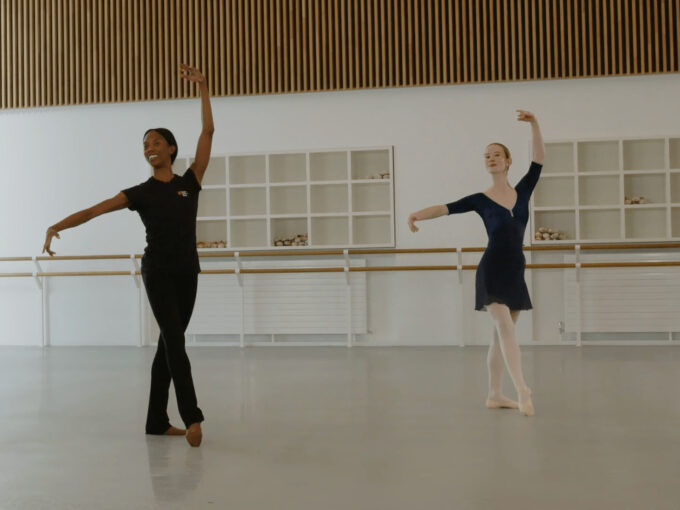 A ballet teacher and a ballet demonstrator are doing a battement tendu at the centre in a studio. The teacher is wearing a black outfit, the demonstrator is wearing a blue leotard and skirt.