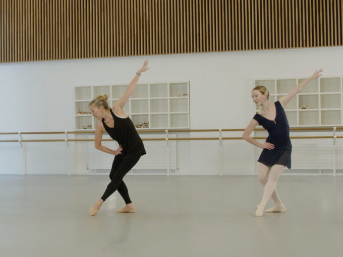 A ballet teacher and a demonstrator are at the centre in a studio. They are in a tendu position, leaning forwards with their back arm raised. The ballet teacher is wearing black clothing, the demonstrator is wearing a blue leotard and skirt.