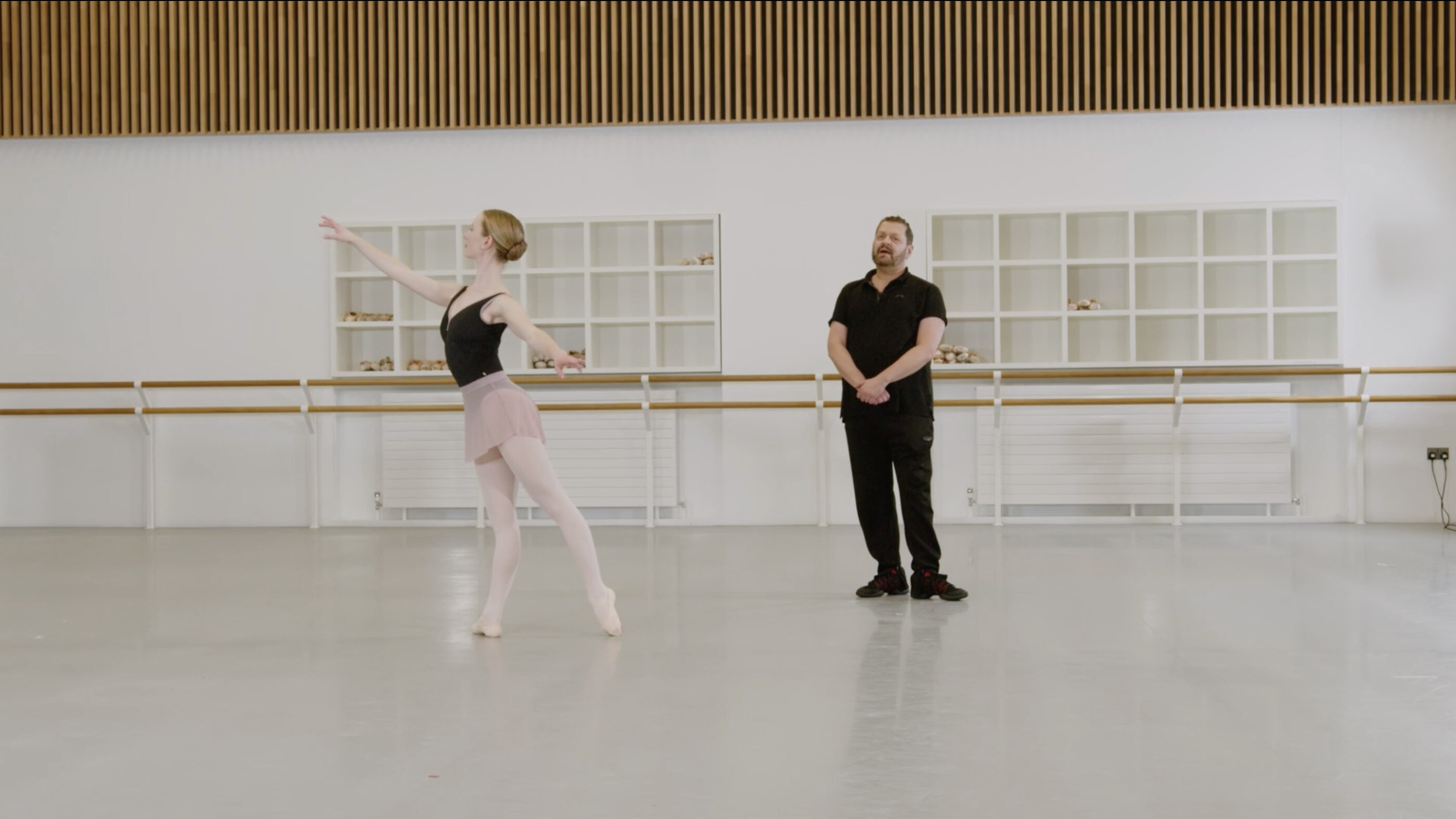 A ballet teacher and a demonstrator are in a dance studio. The demonstrator is doing an arabesque in tendu while the teacher is looking in front of him. The demonstrator is wearing a dark leotard and pink skirt, the teacher is wearing a black t-shirt and trousers.