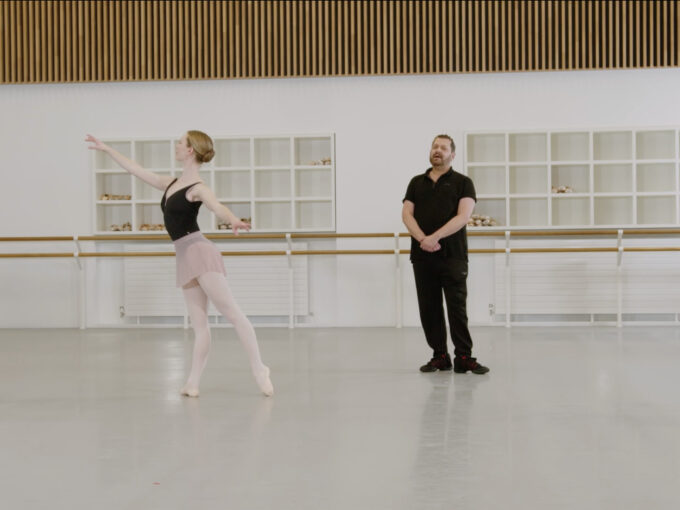 A ballet teacher and a demonstrator are in a dance studio. The demonstrator is doing an arabesque in tendu while the teacher is looking in front of him. The demonstrator is wearing a dark leotard and pink skirt, the teacher is wearing a black t-shirt and trousers.