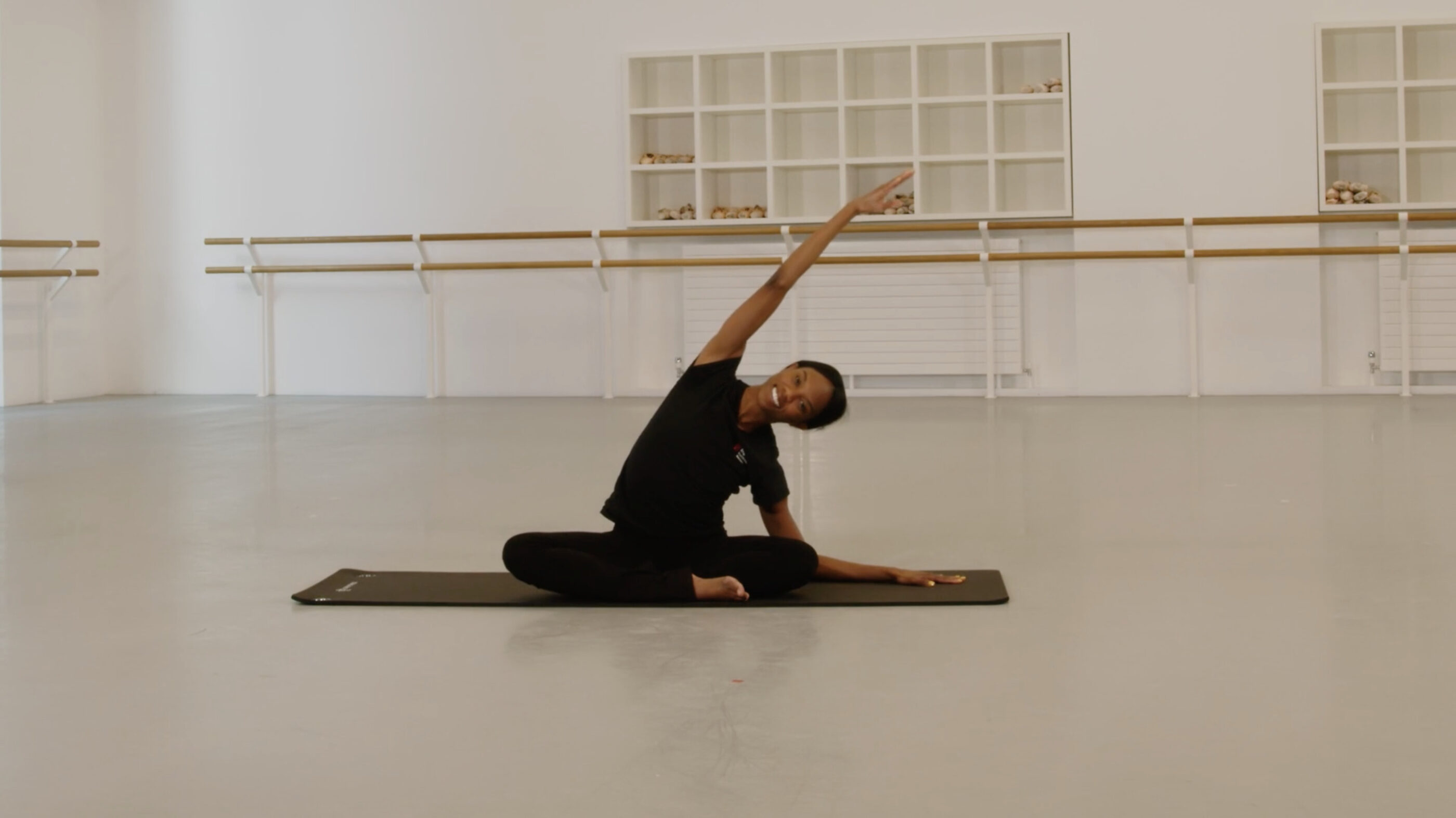 A dance teacher is sitting on a yoga mat in a dance studio. Her legs are folded and she is stretching to the side. She is wearing a black t-shirt and black trousers.