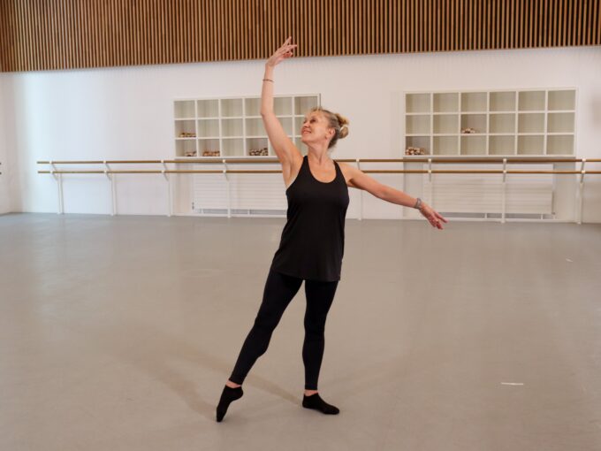 A ballet teacher is at the centre of a dance studio. She's doing a tendu while smiling. She's wearing black clothing.