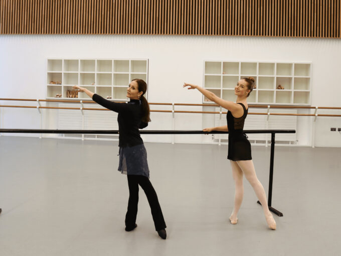 A ballet teacher and a demonstrator are doing a tendu arabesque at the barre. They are in a bright dance studio and are both wearing black clothing.