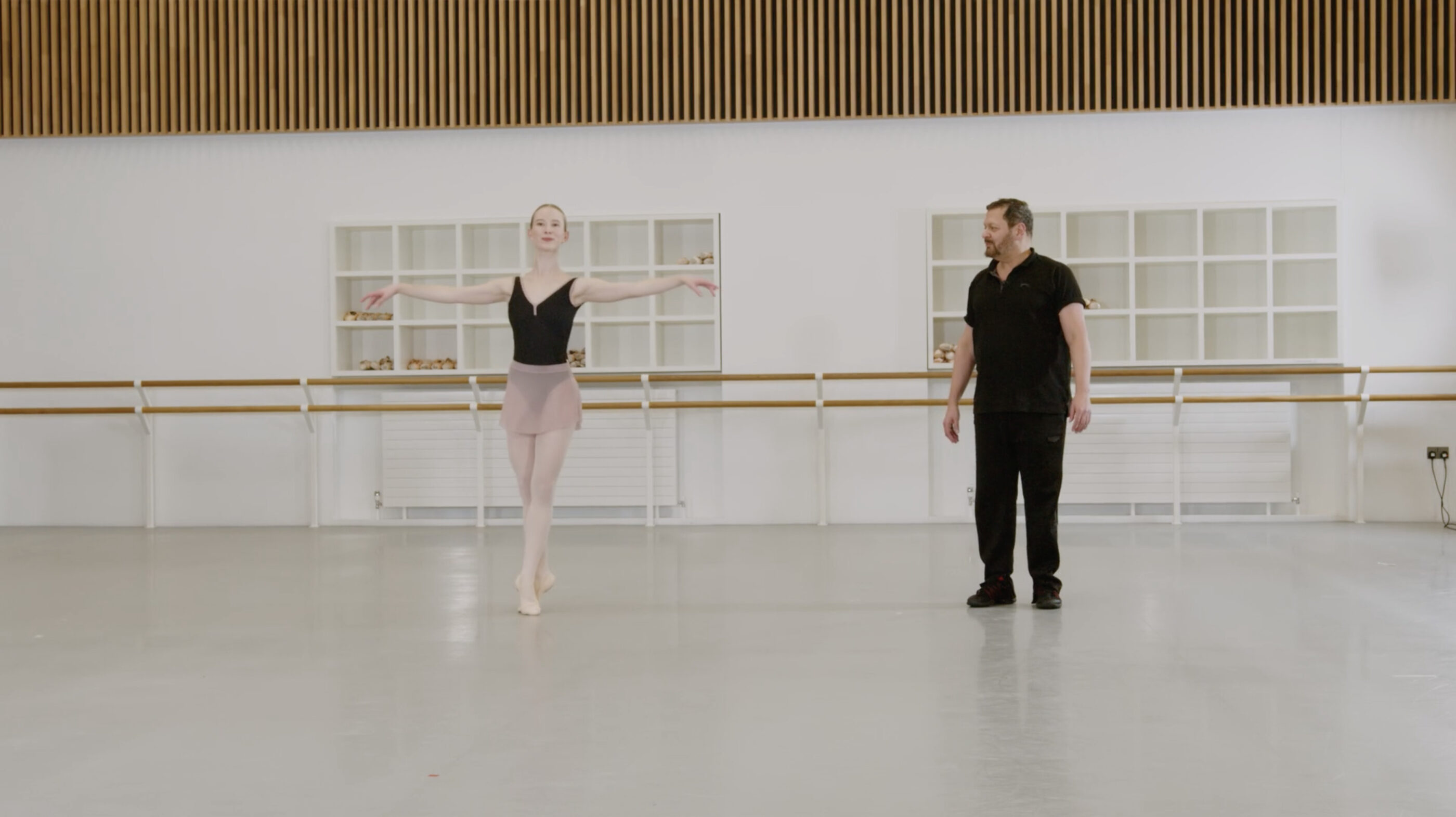 A ballet teacher and a demonstrator are in a bright dance studio. The demonstrator is on her toes while doing a pas de bourree while the teacher is looking towards her. The demonstrator is wearing a black leotard and pink skirt while the teacher is wearing black clothing.