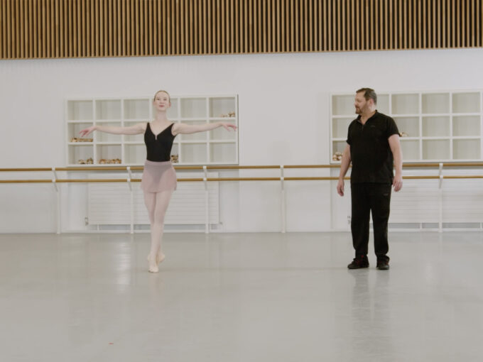 A ballet teacher and a demonstrator are in a bright dance studio. The demonstrator is on her toes while doing a pas de bourree while the teacher is looking towards her. The demonstrator is wearing a black leotard and pink skirt while the teacher is wearing black clothing.